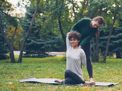 Yoga teacher guiding a student into a seated stretch on a mat outdoors